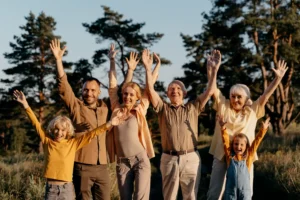 Grupo de personas de diferentes edades, incluyendo niños y adultos mayores, levantando las manos en señal de celebración al aire libre, rodeados de árboles y naturaleza.
