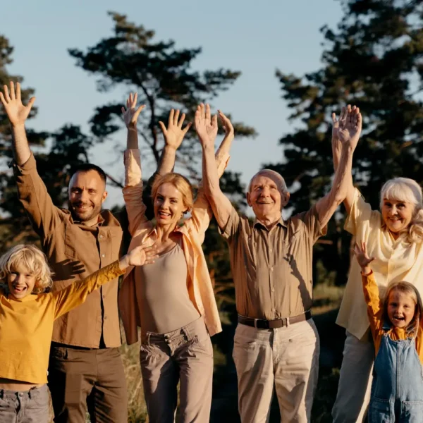 Grupo de personas de diferentes edades, incluyendo niños y adultos mayores, levantando las manos en señal de celebración al aire libre, rodeados de árboles y naturaleza.