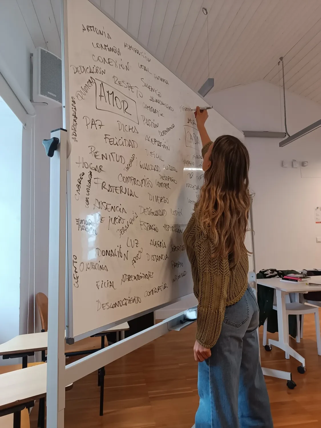 Mujer escribiendo en una pizarra blanca con palabras relacionadas con el amor y las relaciones, en un ambiente de aula. Ella viste un suéter de punto y jeans.