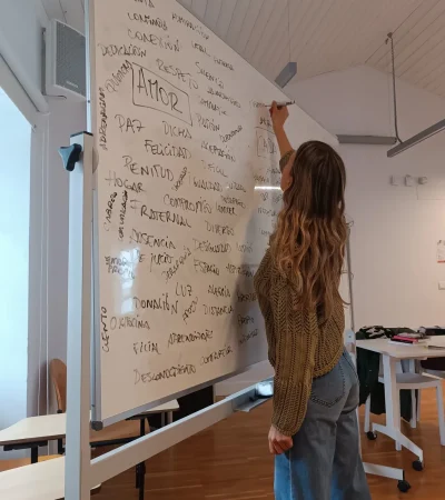 Mujer escribiendo en una pizarra blanca con palabras relacionadas con el amor y las relaciones, en un ambiente de aula. Ella viste un suéter de punto y jeans.